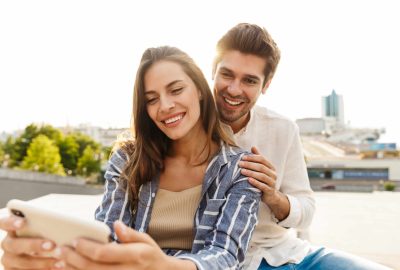 Image of young caucasian happy couple smiling and hugging while sitting outdoors with cellphone